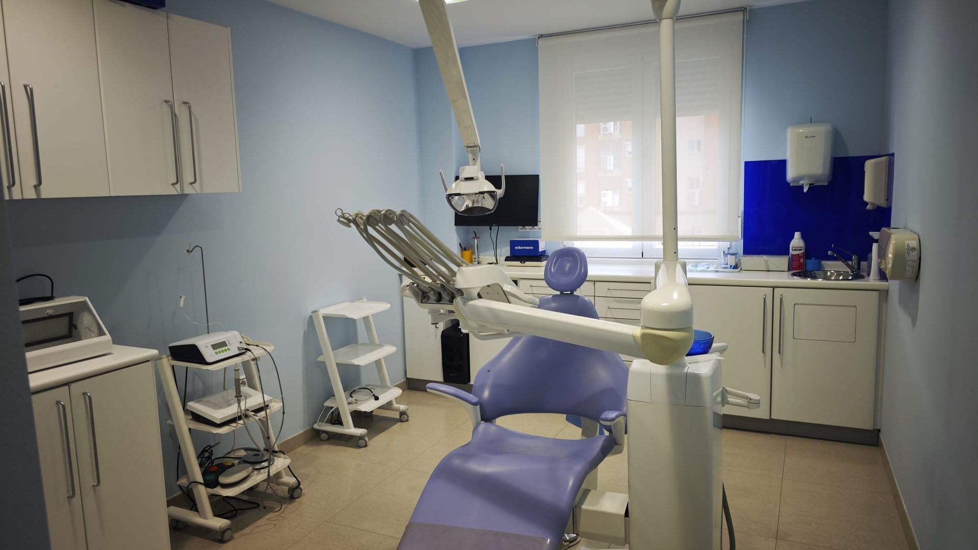 Sala de examen dental con sillón, luz cenital, armarios y equipo médico en una clínica azul y blanca.