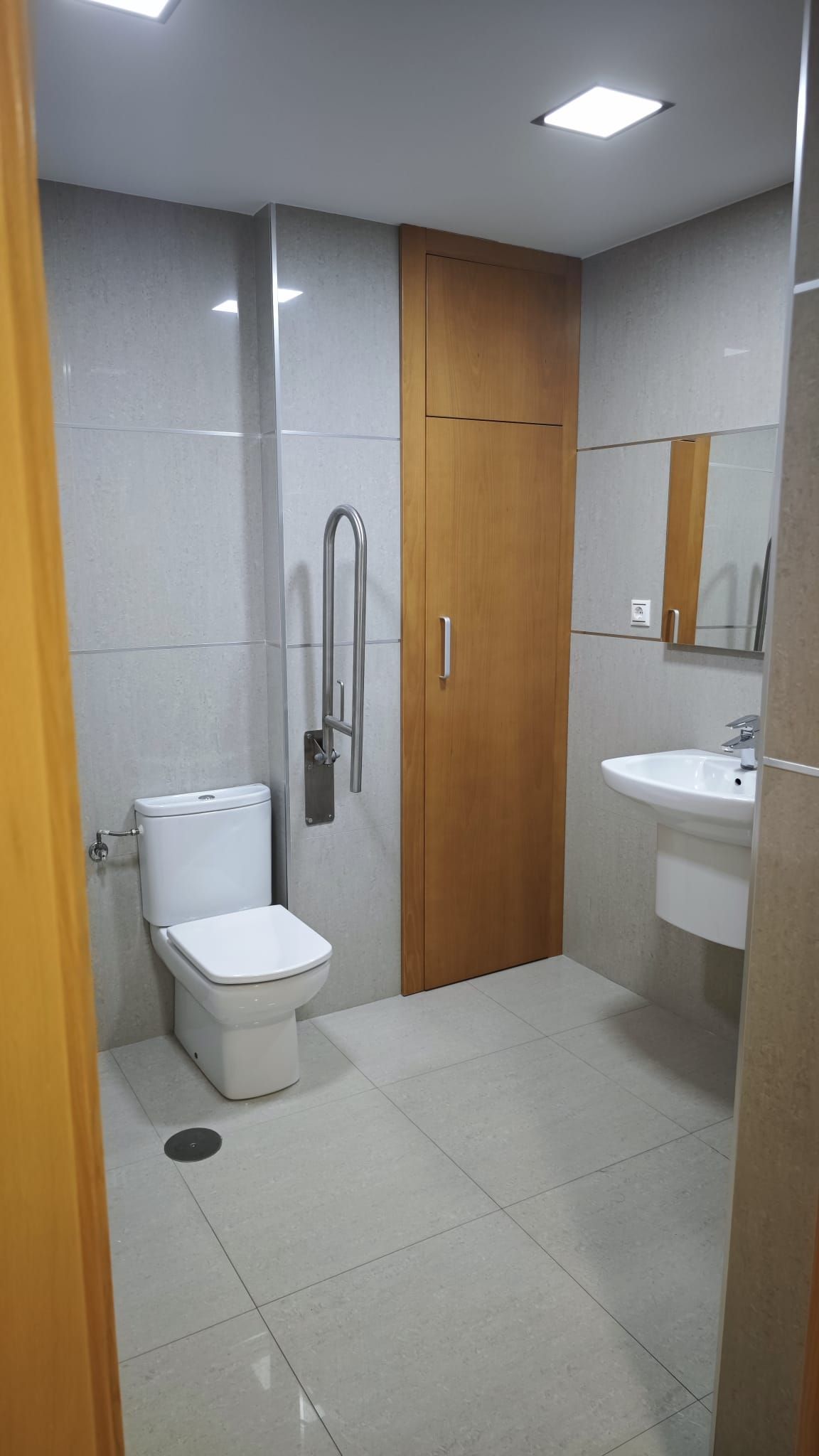 Baño accesible con inodoro, ducha, lavabo y puerta de madera en el suelo de baldosas claras.