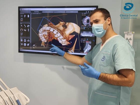 Sala de examen dental con sillón, luz cenital, armarios y equipo médico en una clínica azul y blanca.
