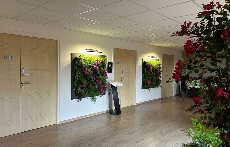 Couloir avec portes beiges et jardinières murales de fleurs vertes et roses, plus une grande plante en pot sur la droite.