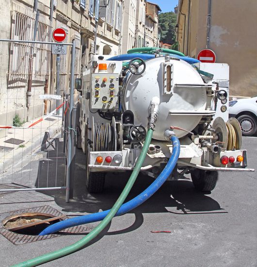 Camion de nettoyage des rues avec des tuyaux bleus et verts sur une rue urbaine étroite, près d'immeubles et de panneaux d'interdiction d'accès.