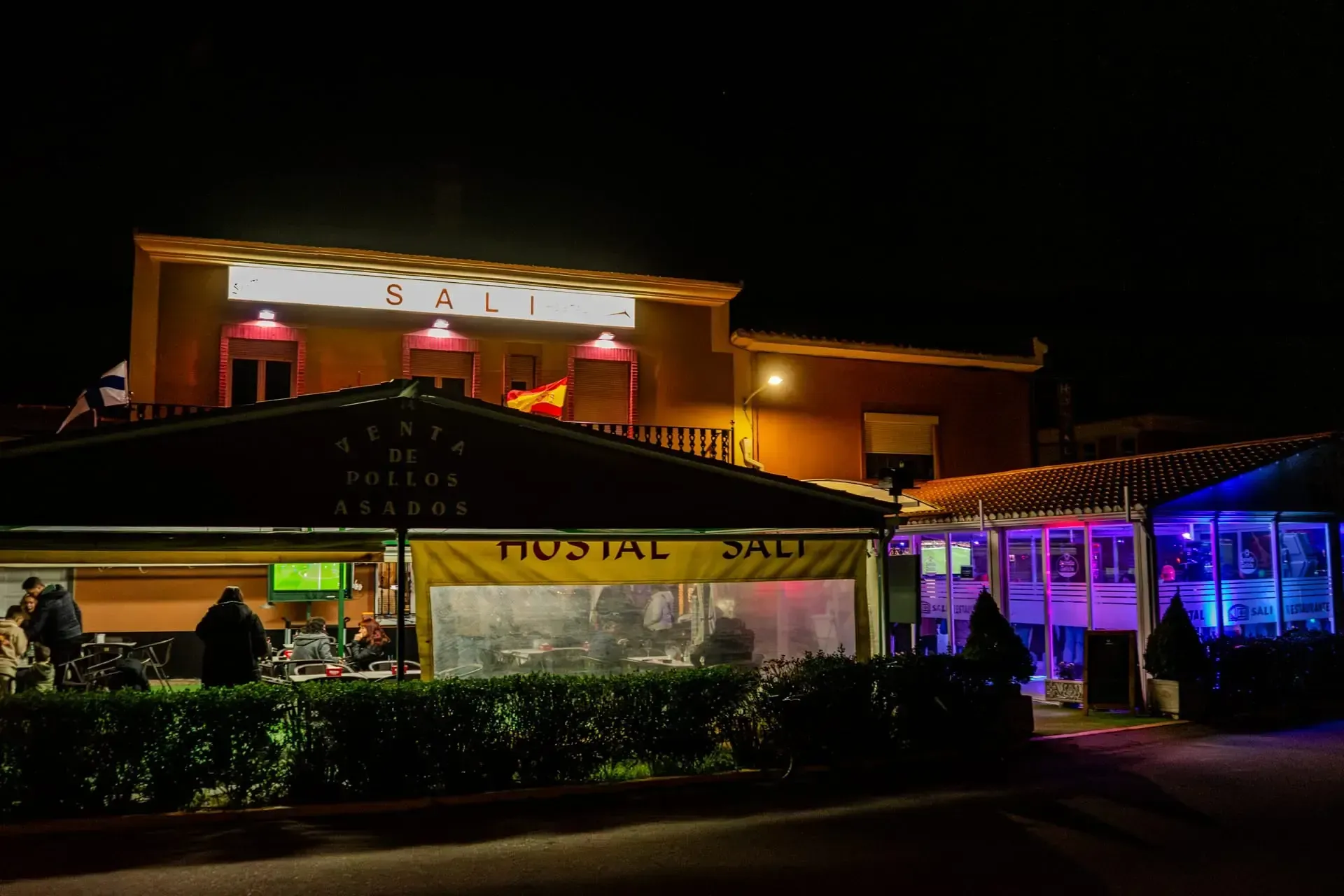 Vista nocturna del exterior iluminado de un hotel con una entrada de cristal y luces de acento de color azul violáceo.
