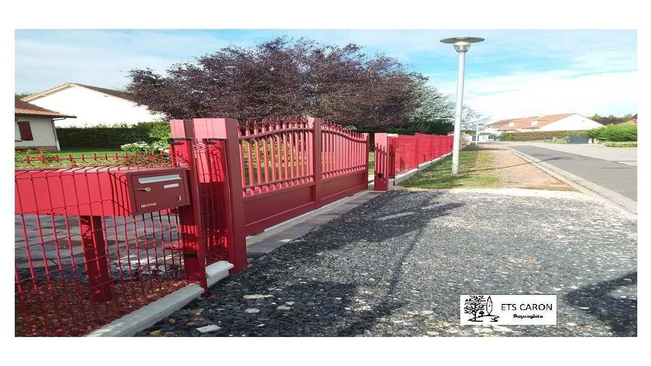 Une clôture rouge borde un chemin de gravier, des maisons blanches et un lampadaire éclaire le ciel.