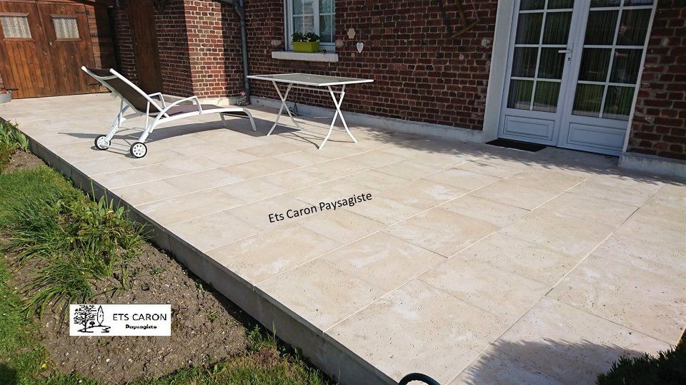 Terrasse avec dalles de pierre couleur crème, deux chaises longues blanches et une petite table à côté d'une maison en briques.