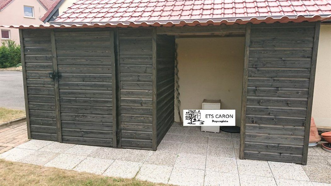 Abri en bois avec volets sombres et toit de tuiles, porte centrale ouverte et petite pancarte devant.