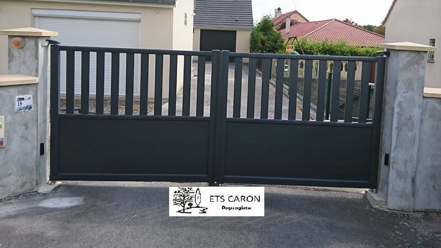Portail coulissant en métal noir entre des piliers en béton devant une maison, avec un petit panneau à sa base.