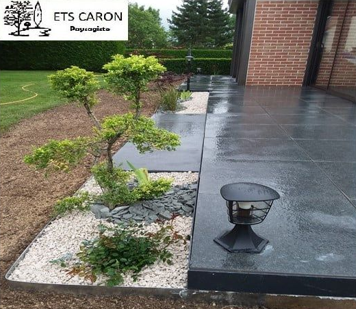 Terrasse moderne en béton avec lampe extérieure noire, dalles de jardin et petits arbustes à côté d'une maison en briques.