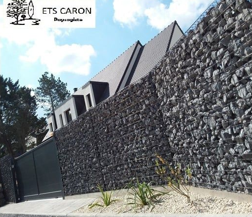 Mur en gabion et bâtiment moderne avec un portail noir sous un ciel bleu.