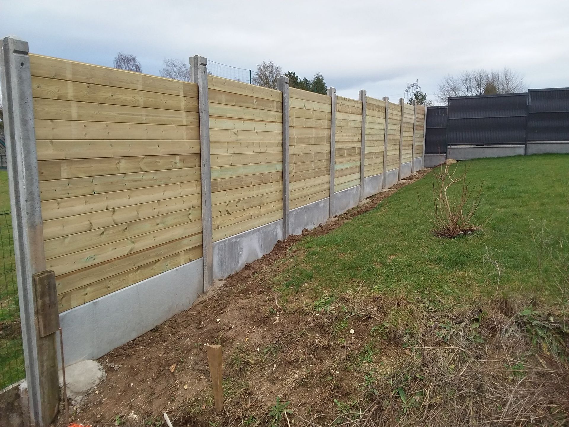 Clôture en bois avec poteaux en béton, préservant l'intimité d'une cour gazonnée sous un ciel couvert.
