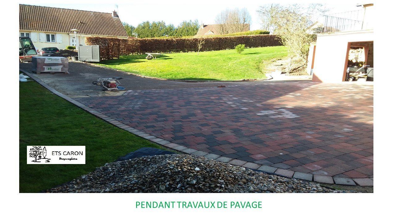 Allée et cour pavées de briques à côté d'une maison, avec un jardin gazonné et un garage ouvert.