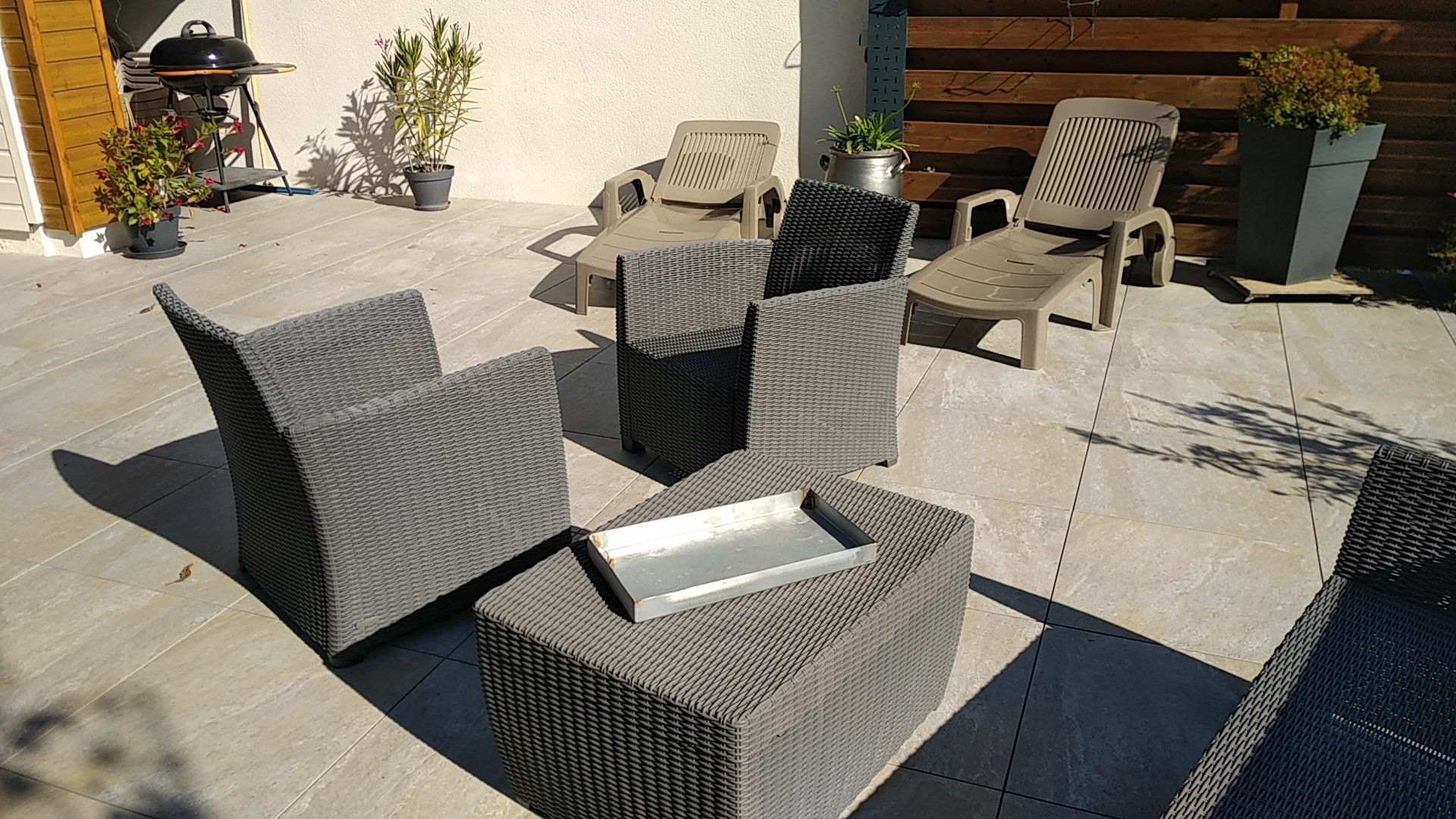 Patio avec chaises en osier gris et petite table sur une terrasse en pierre ensoleillée.
