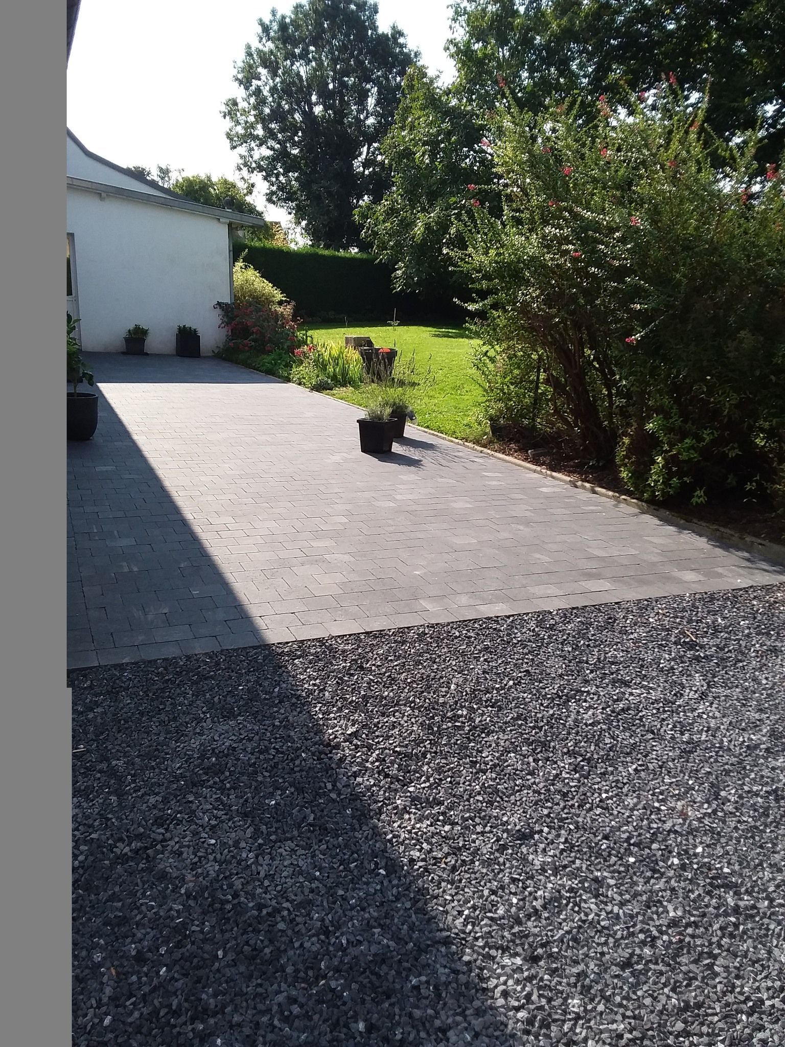 Terrasse en gravier jouxtant une maison blanche, avec des buissons et des arbres baignés de soleil.