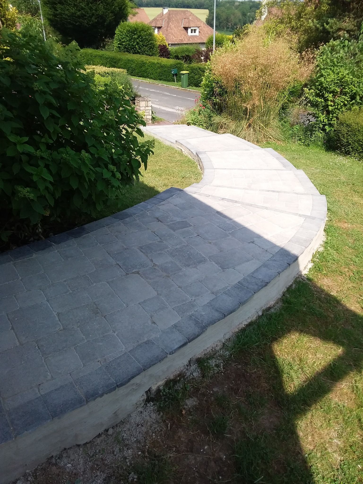 Allée pavée incurvée longeant des haies vertes, traversant un quartier résidentiel ensoleillé.