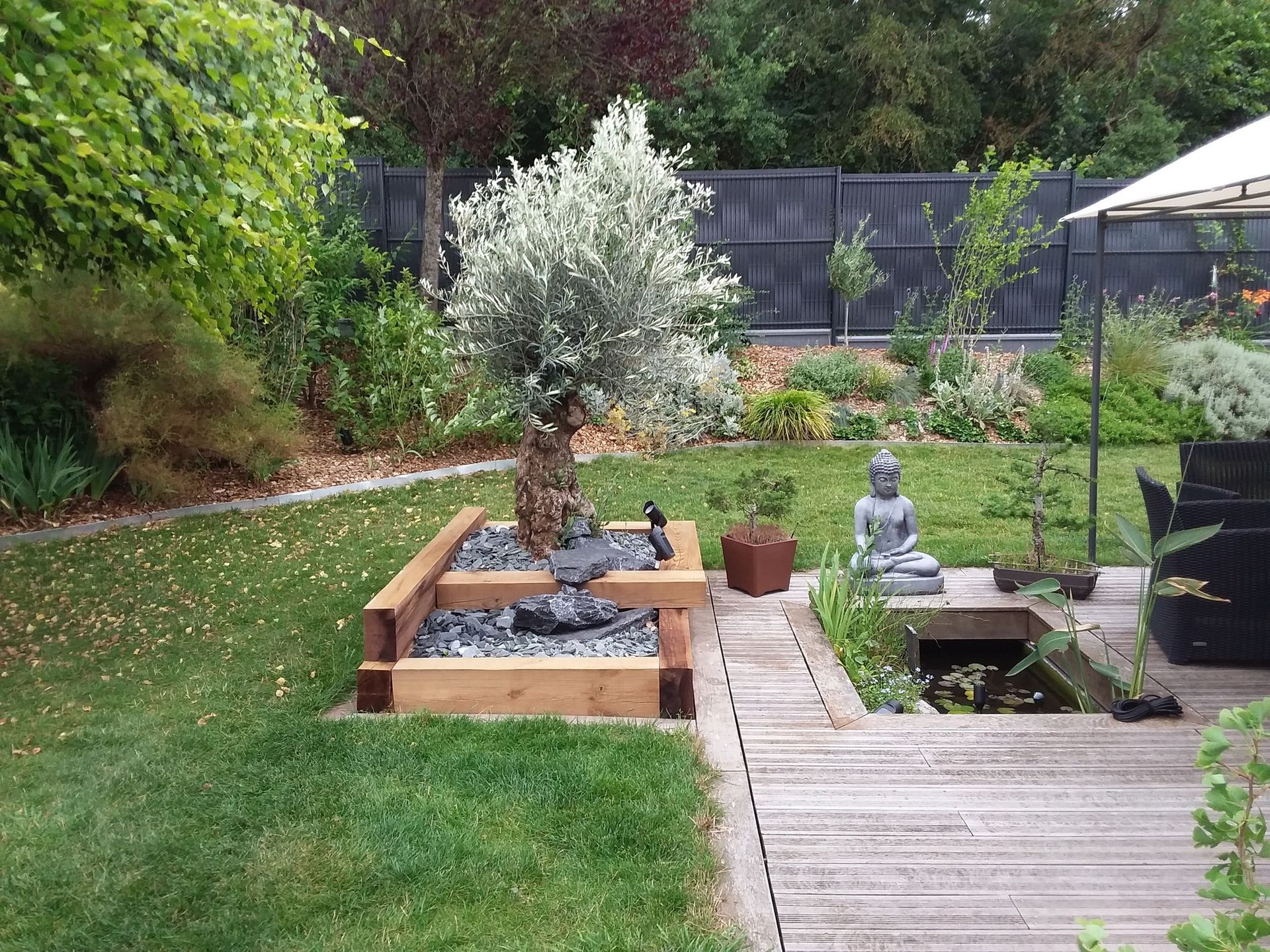 Jardin arrière avec un petit étang, un arbre en pot, une terrasse en bois et une statue de Bouddha assis.