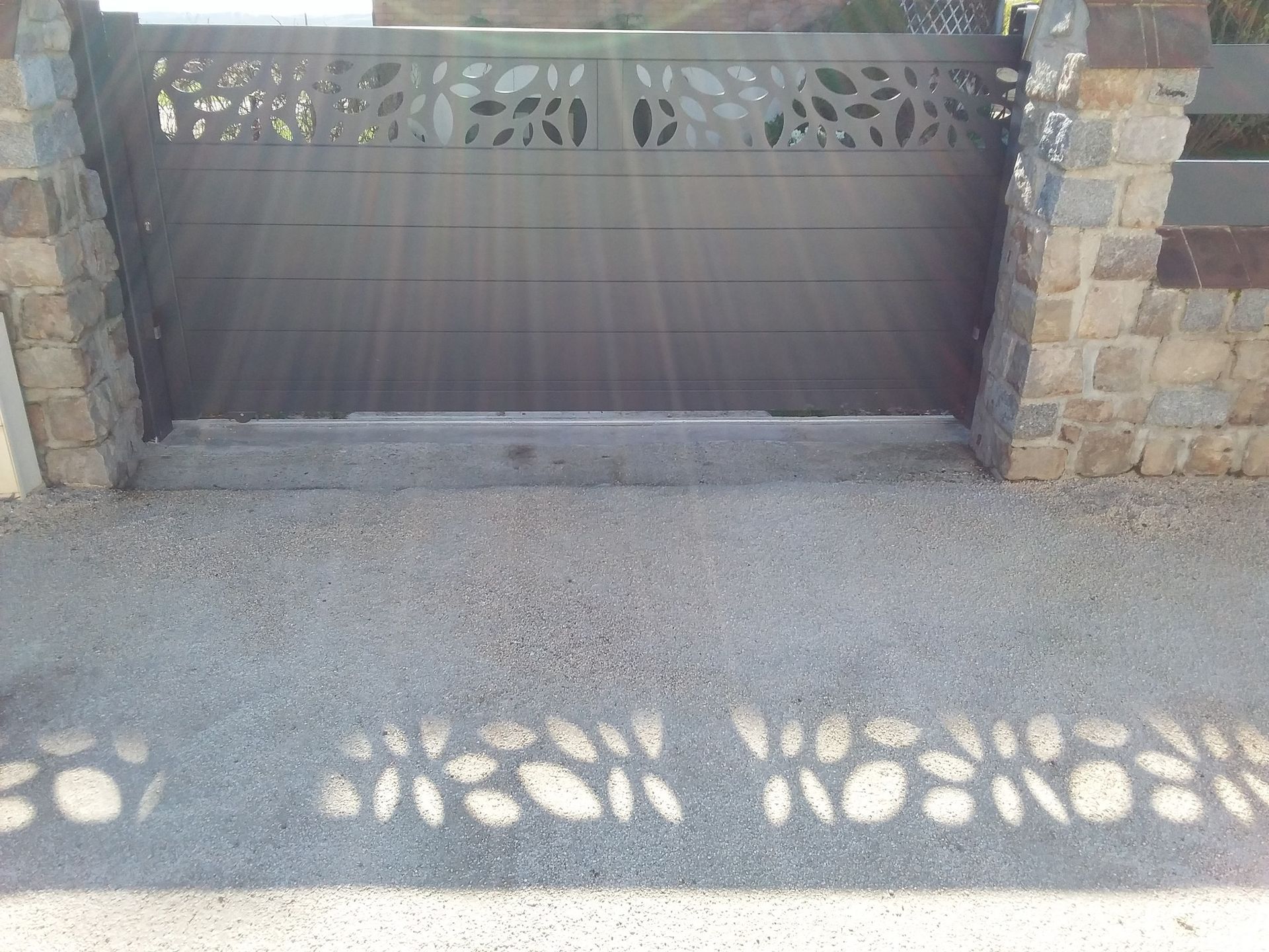 Portail d'allée en pierre avec un panneau en métal noir et des jeux d'ombres et de lumière sur le pavé.