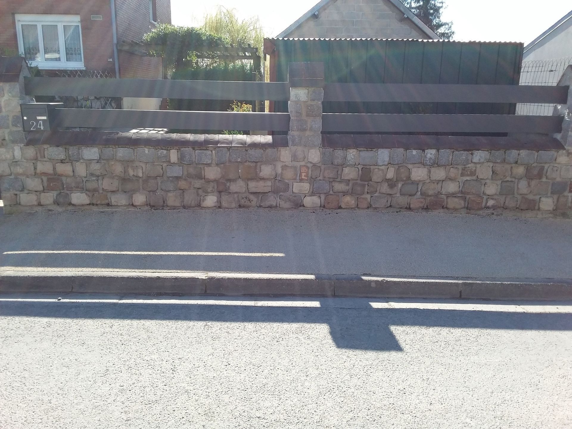 Mur en pierre et clôture en bois le long d'une route, avec une maison en arrière-plan et de longues ombres sur le trottoir.