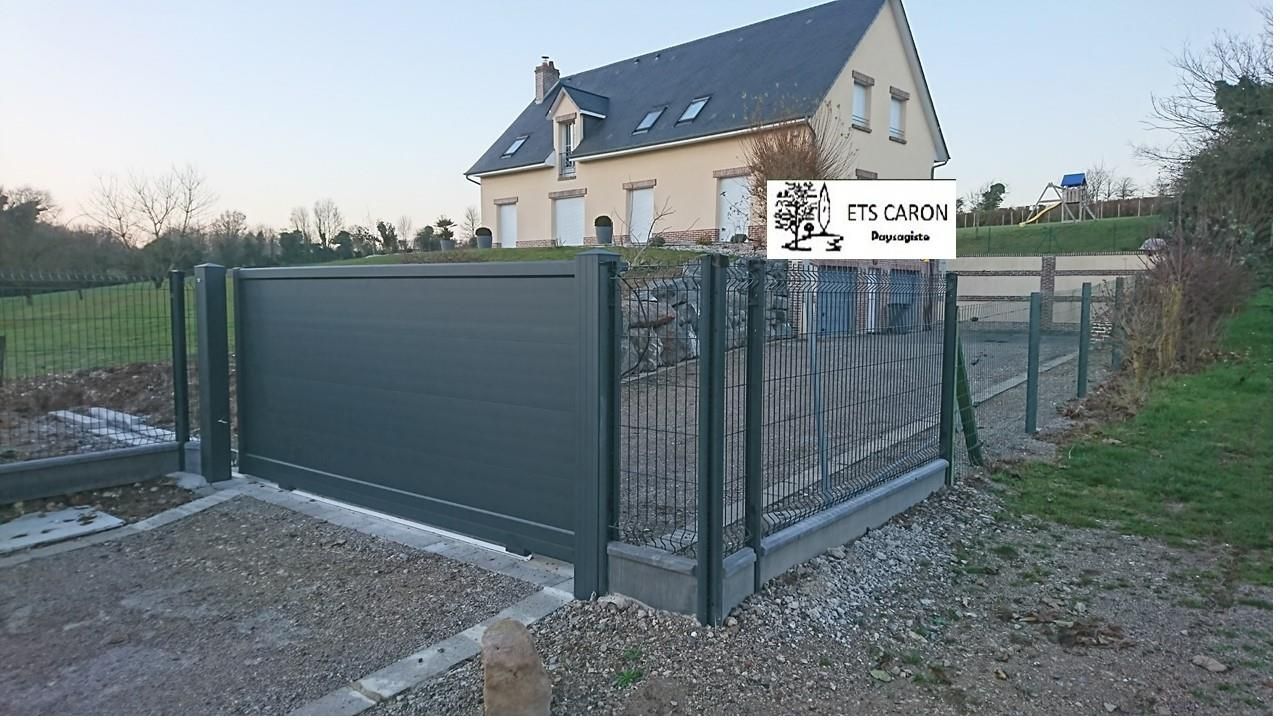 Maison moderne avec clôture et portail sombres, donnant sur une allée de gravier dans une cour rurale.