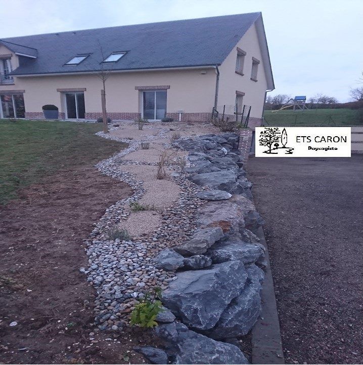 Tranchée de drainage bordée de pierres à côté d'une maison beige avec un toit en pente et un jardin.