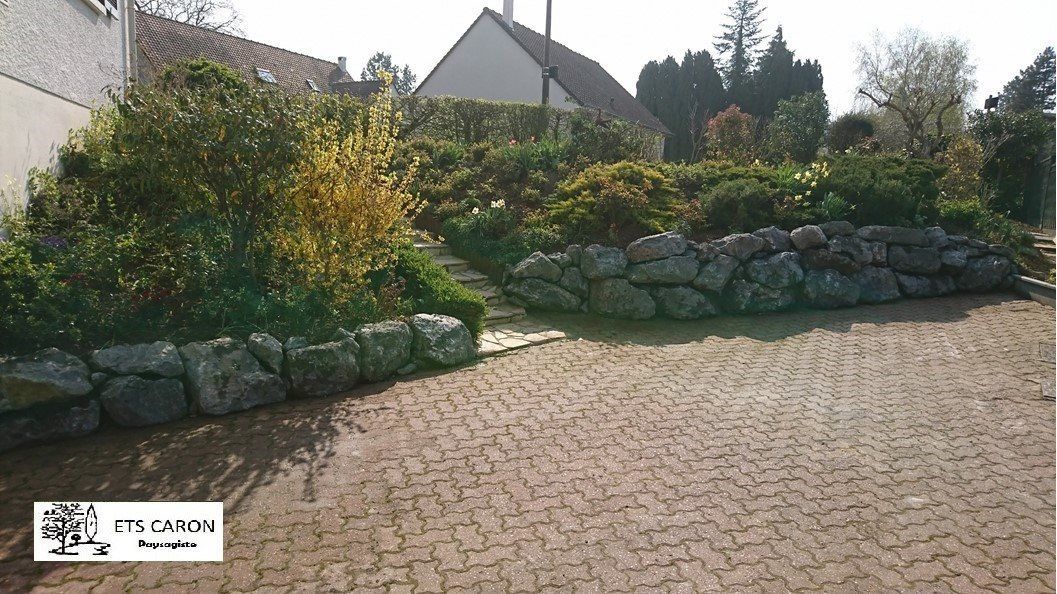 Allée en pierre longeant un jardin paysager avec arbustes, fleurs et maisons en arrière-plan.