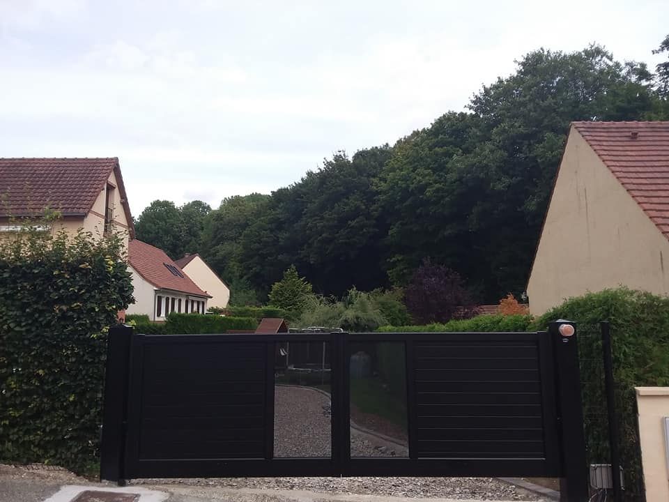 Portail d'allée noir devant une maison de banlieue, avec des haies et des arbres derrière.