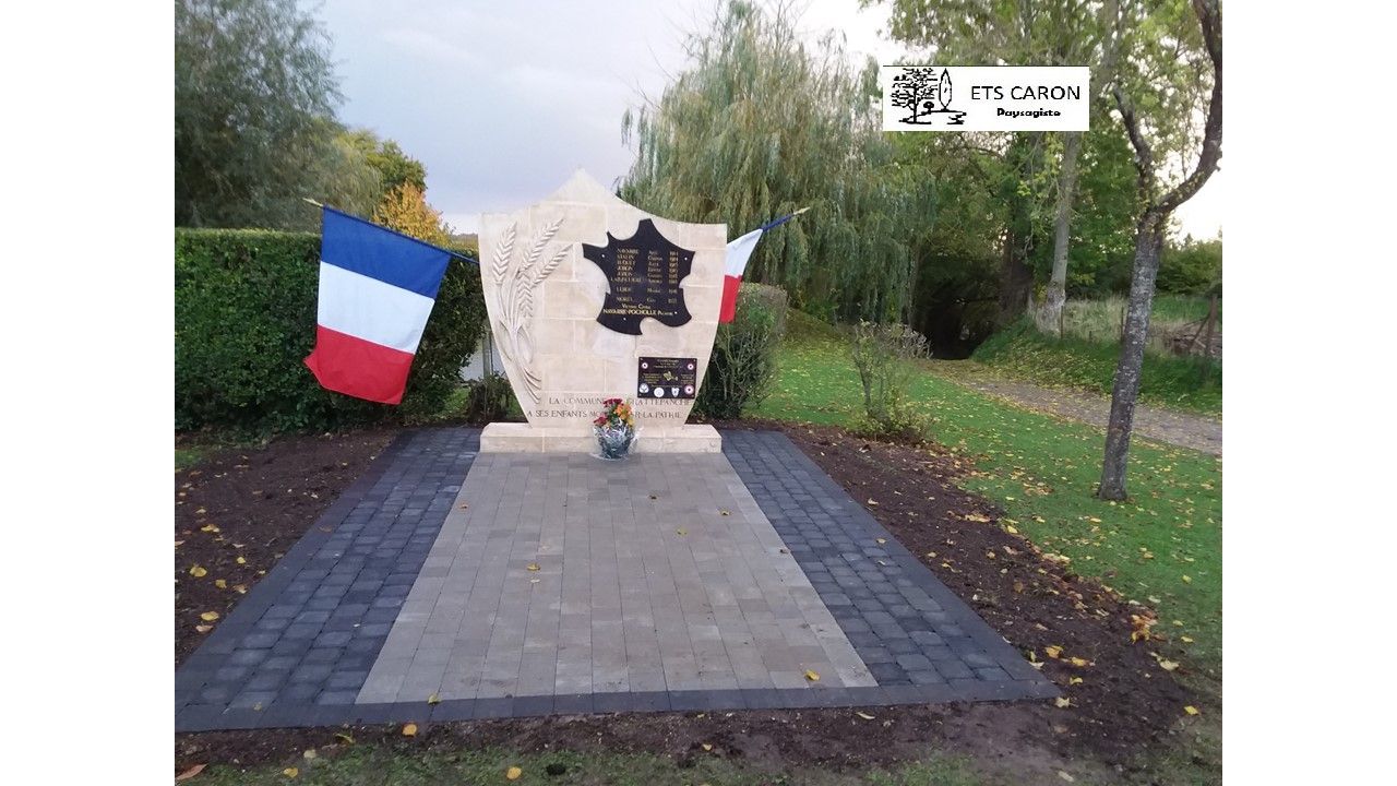 Plaque commémorative dans un jardin orné de drapeaux français et blancs, d'une couronne et d'une allée pavée.