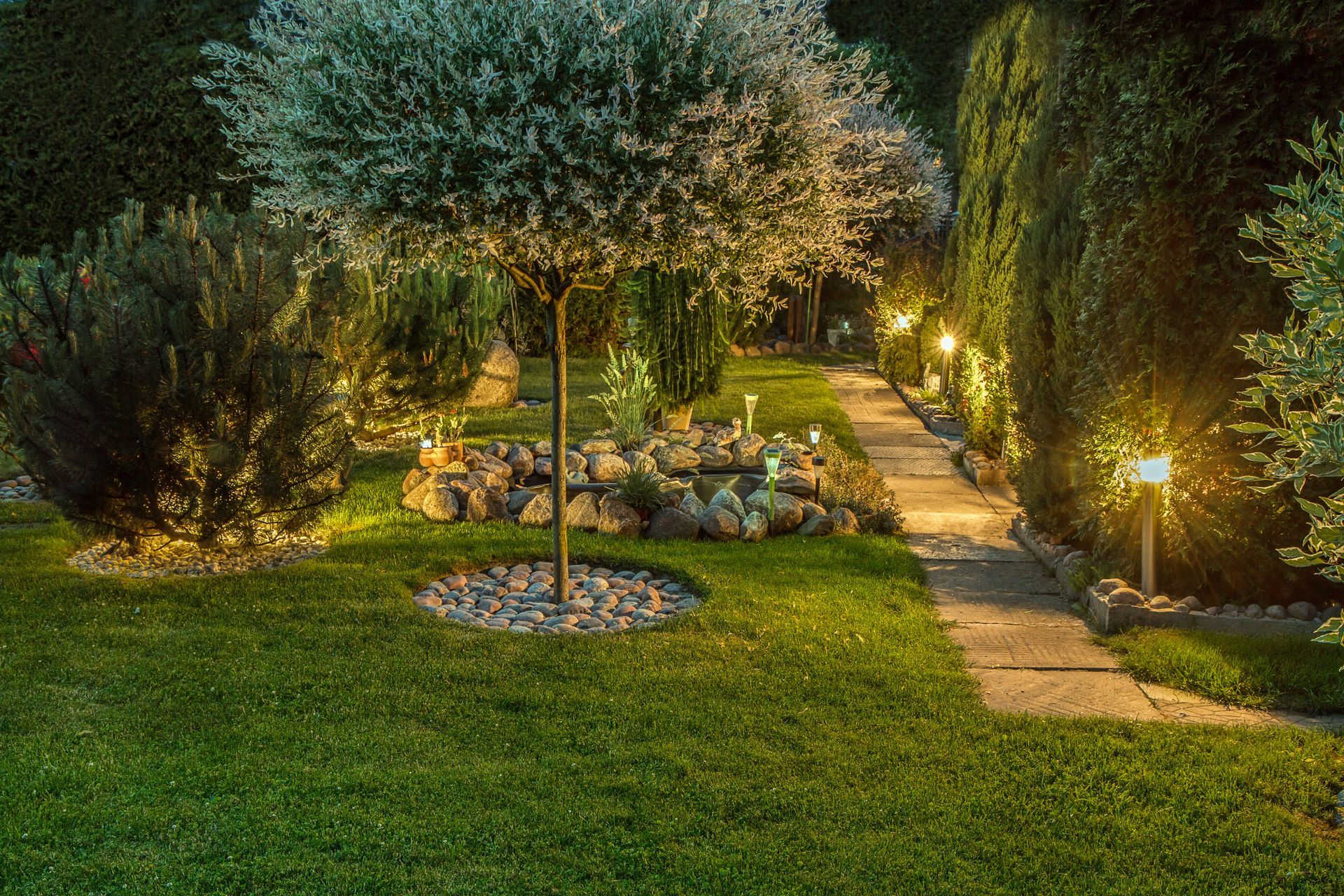 Au crépuscule, un chemin de jardin éclairé longe une pelouse et un arbre, tandis que des lumières chaudes illuminent arbustes et fleurs.