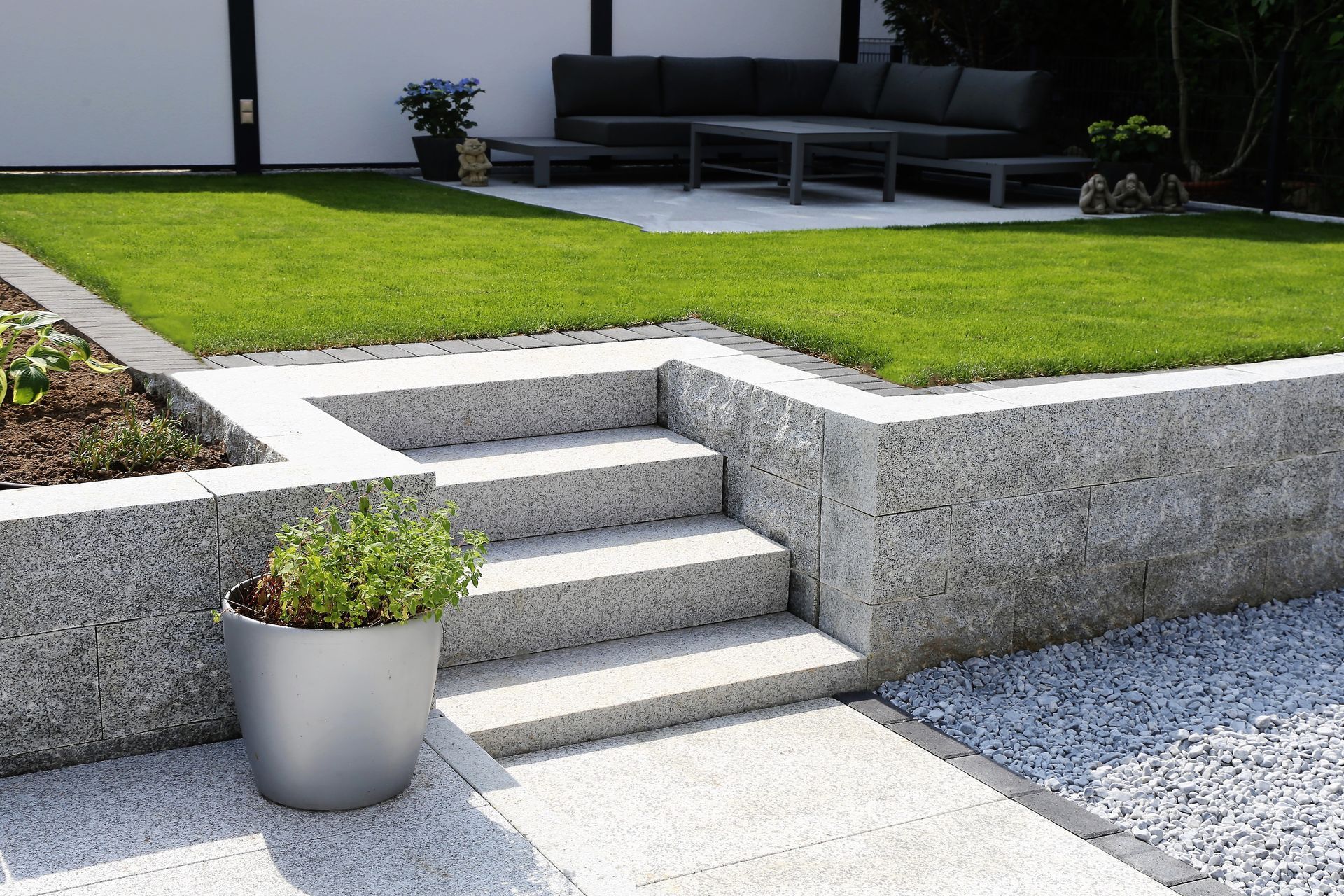 Terrasse moderne avec murs de soutènement en pierre grise, pelouse verte et plante en pot près de l'escalier.