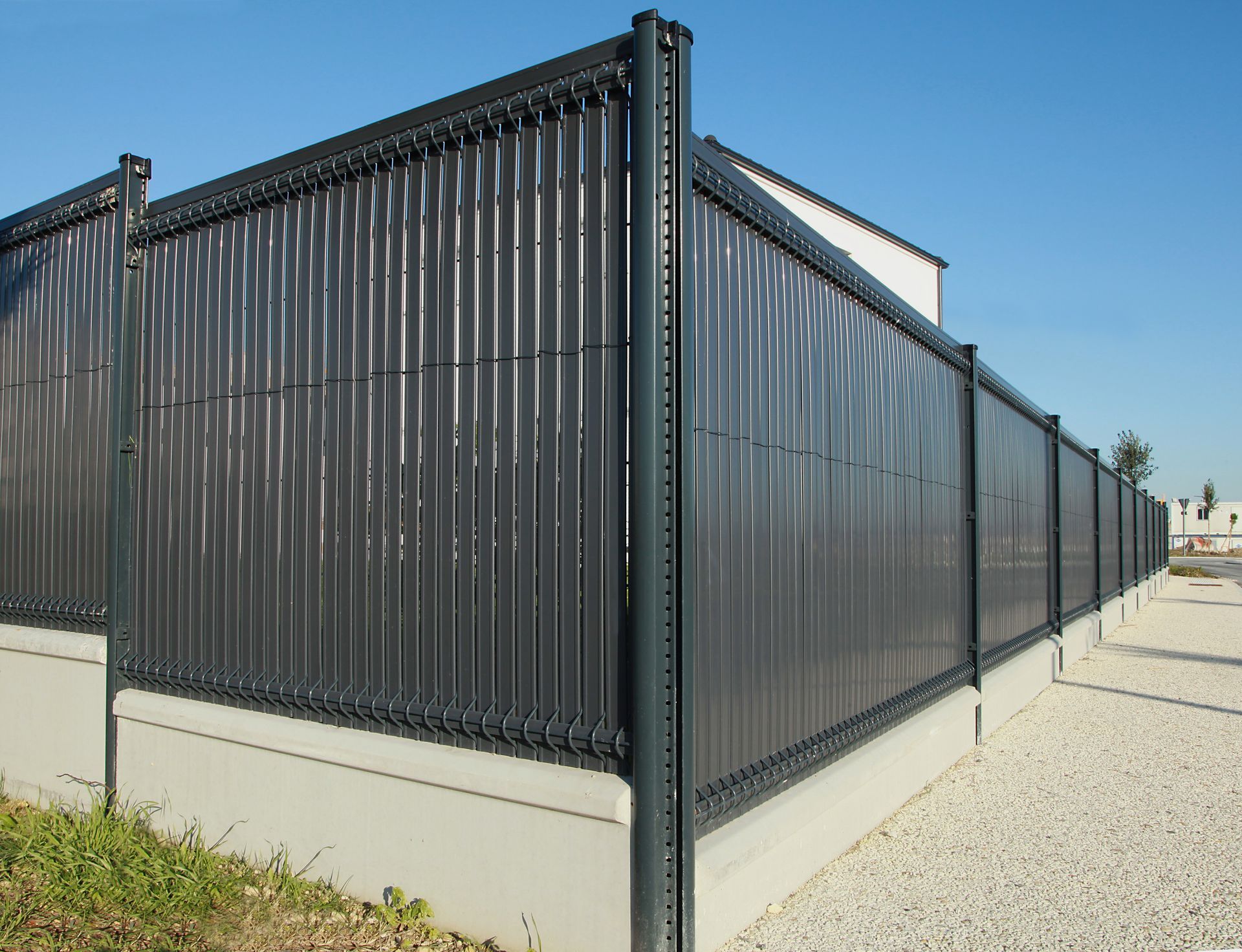Clôture métallique noire assurant l'intimité le long d'un mur clair dans une zone industrielle, vue depuis un trottoir.