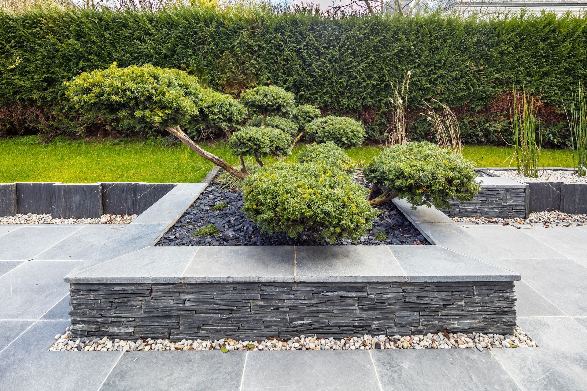 Jardin paysager moderne avec arbustes taillés, parterres de gravier foncé et jardinière basse en pierre devant une haie.