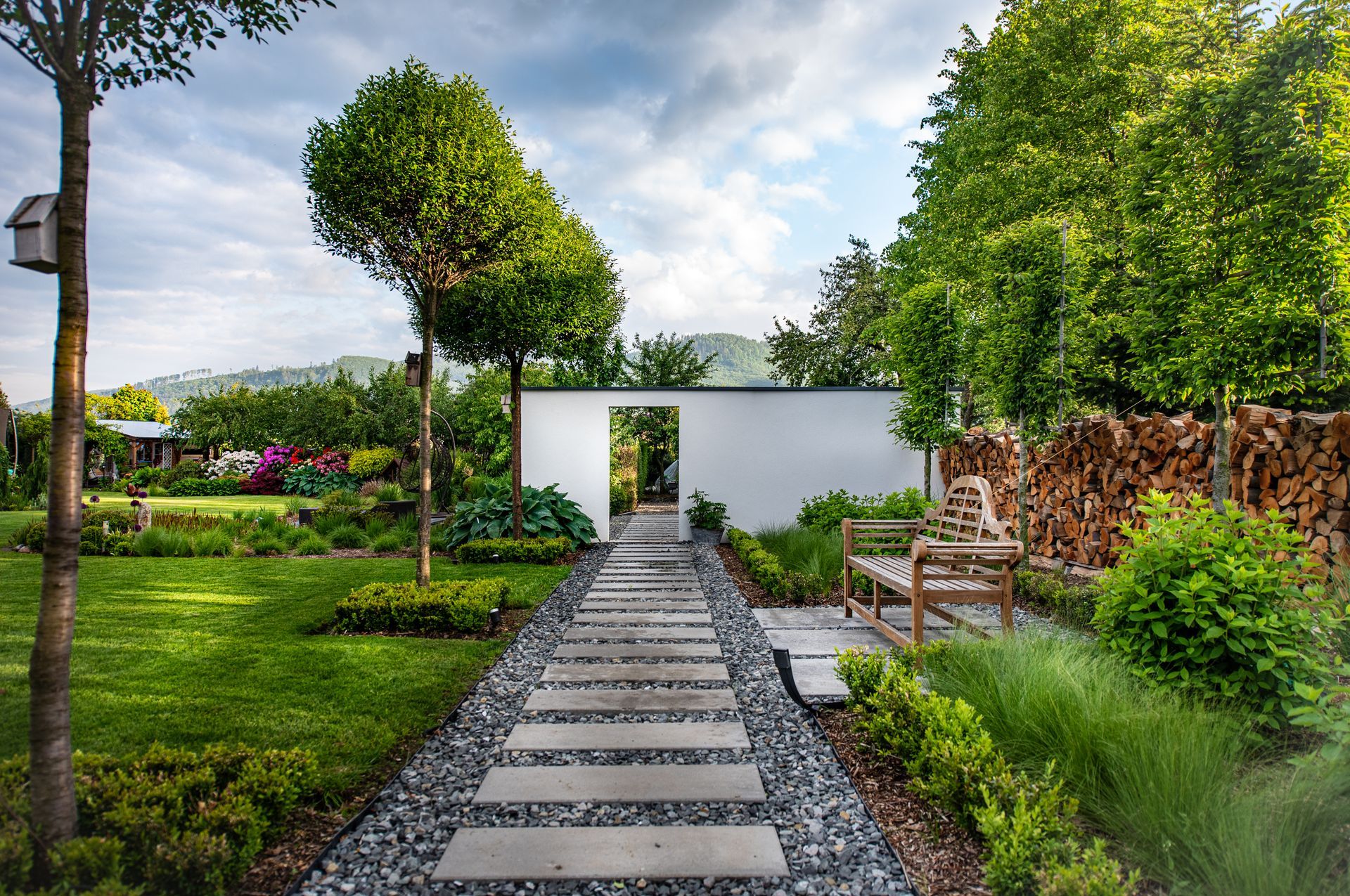 Allée de jardin paysager moderne avec des dalles de pierre menant à un mur blanc et un portail, entourée d'une végétation luxuriante.