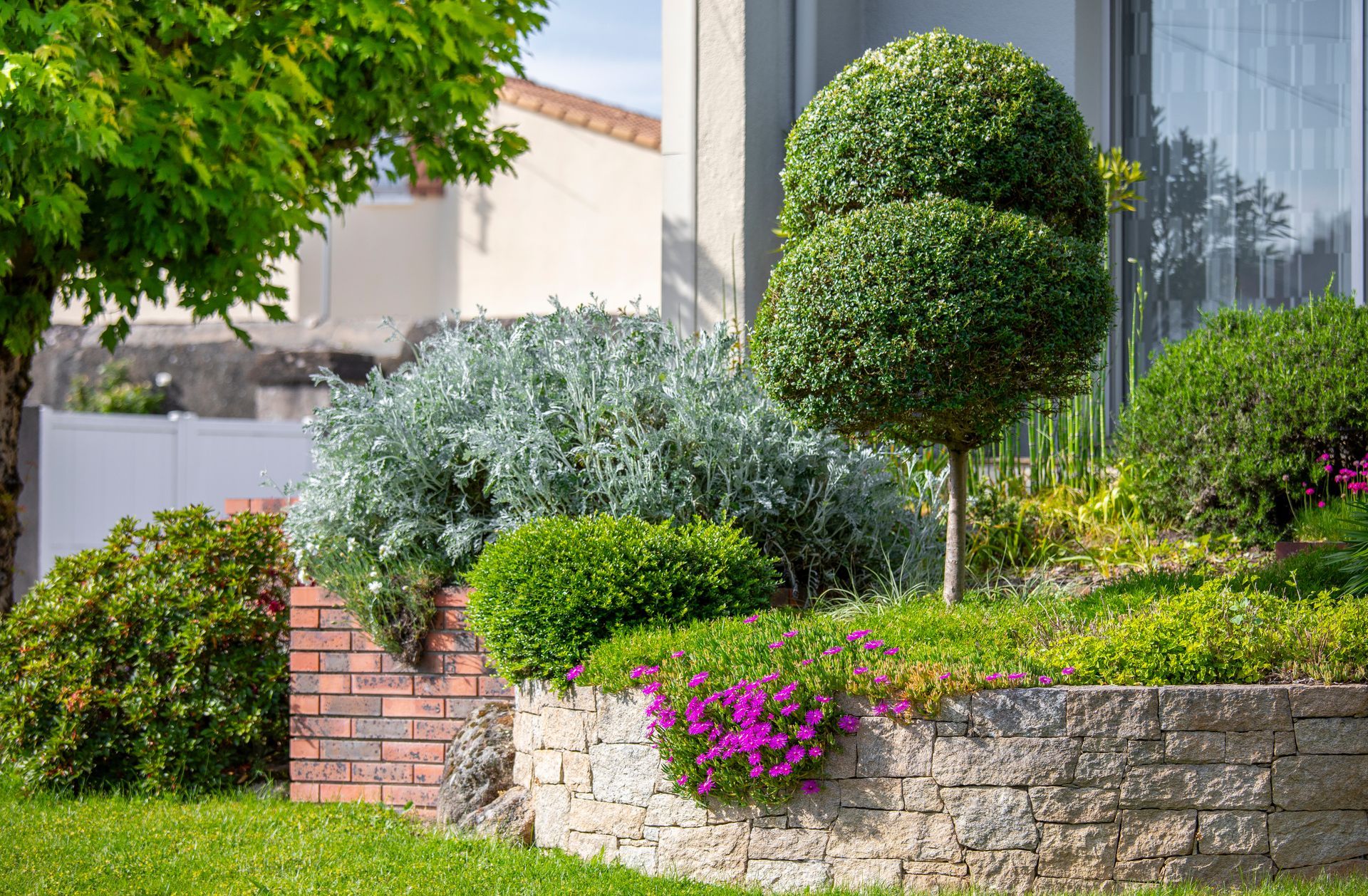 Jardin paysager avec arbustes taillés, mur de soutènement en pierre et fleurs roses à côté d'une maison.