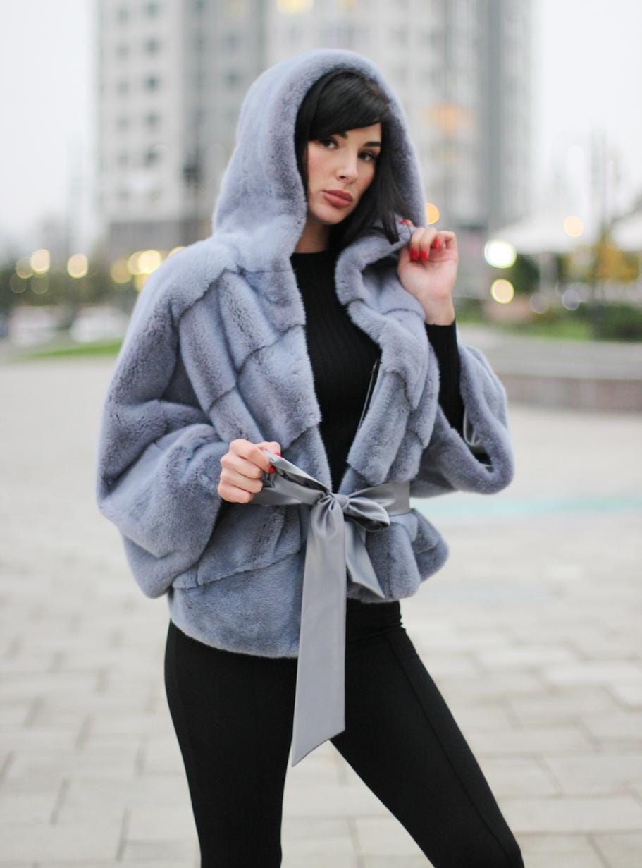 Mujer con abrigo de piel azul con capucha, top negro y leggings atados a la cintura, de pie en el exterior.