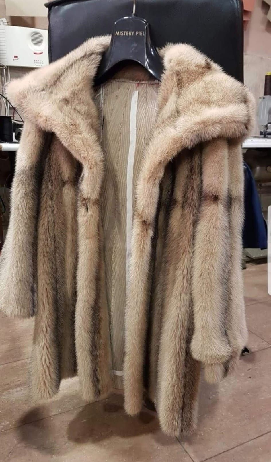 Un abrigo de piel color canela con capucha, colgado en una percha negra, sobre un fondo borroso.