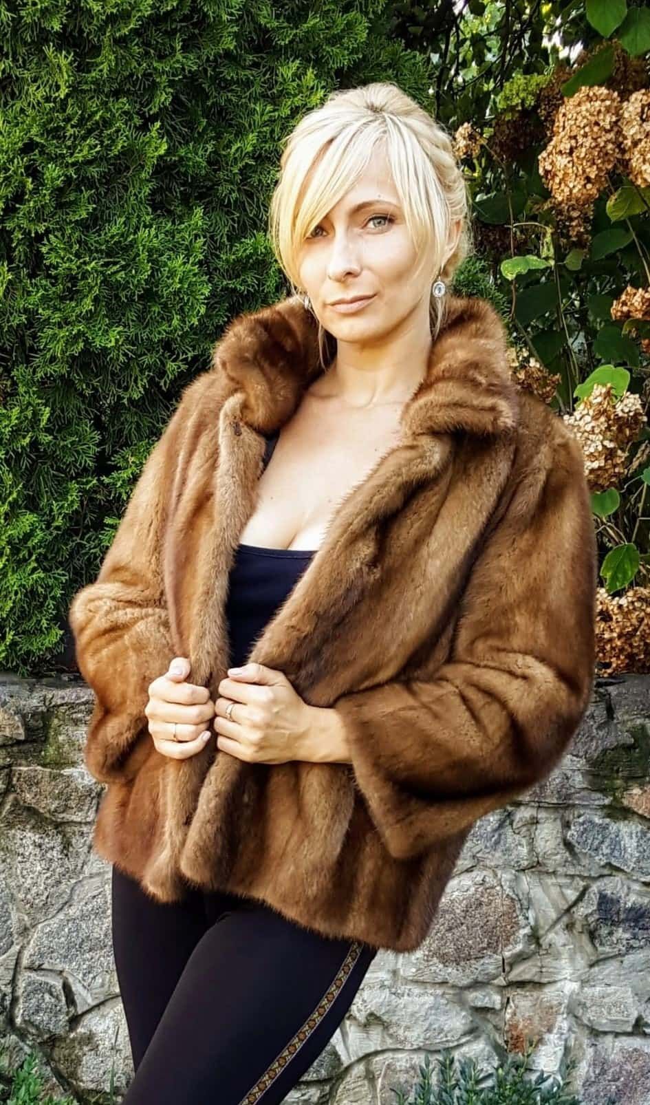 Mujer con abrigo de piel marrón, que sujeta cerrado, con cabello rubio, con fondo de vegetación y pared de piedra.