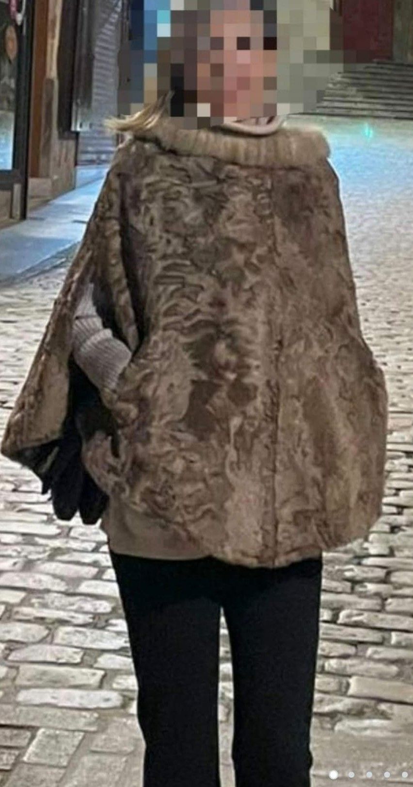 Persona con capa de piel marrón con bolsillos, pantalones negros y guantes, de pie en una calle empedrada por la noche.