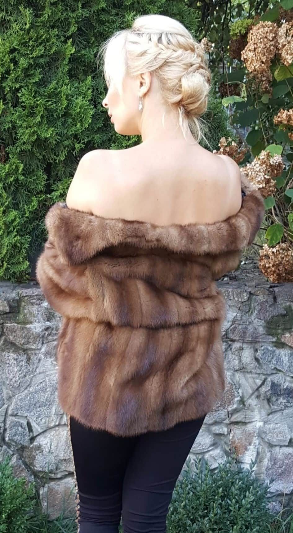 Mujer con chaqueta de piel marrón de hombros descubiertos, de espaldas al espectador, de pie al aire libre.