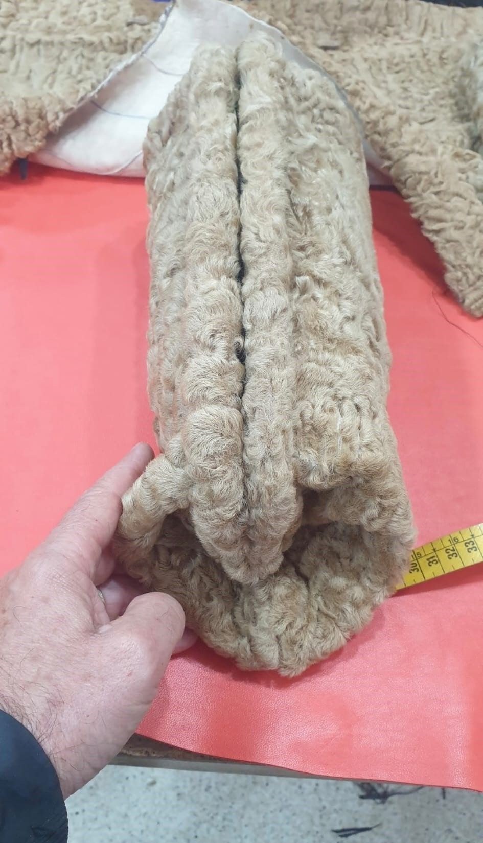 La mano de una persona sostiene una tela peluda de color marrón claro, posiblemente parte de un animal de peluche, sobre una superficie roja.