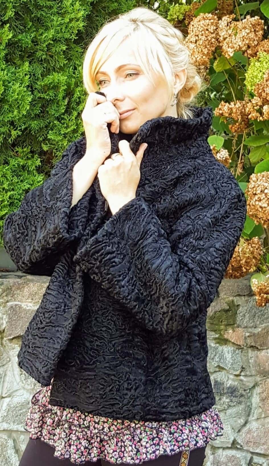 Mujer con abrigo negro texturizado, manos en el cuello, de pie al aire libre.