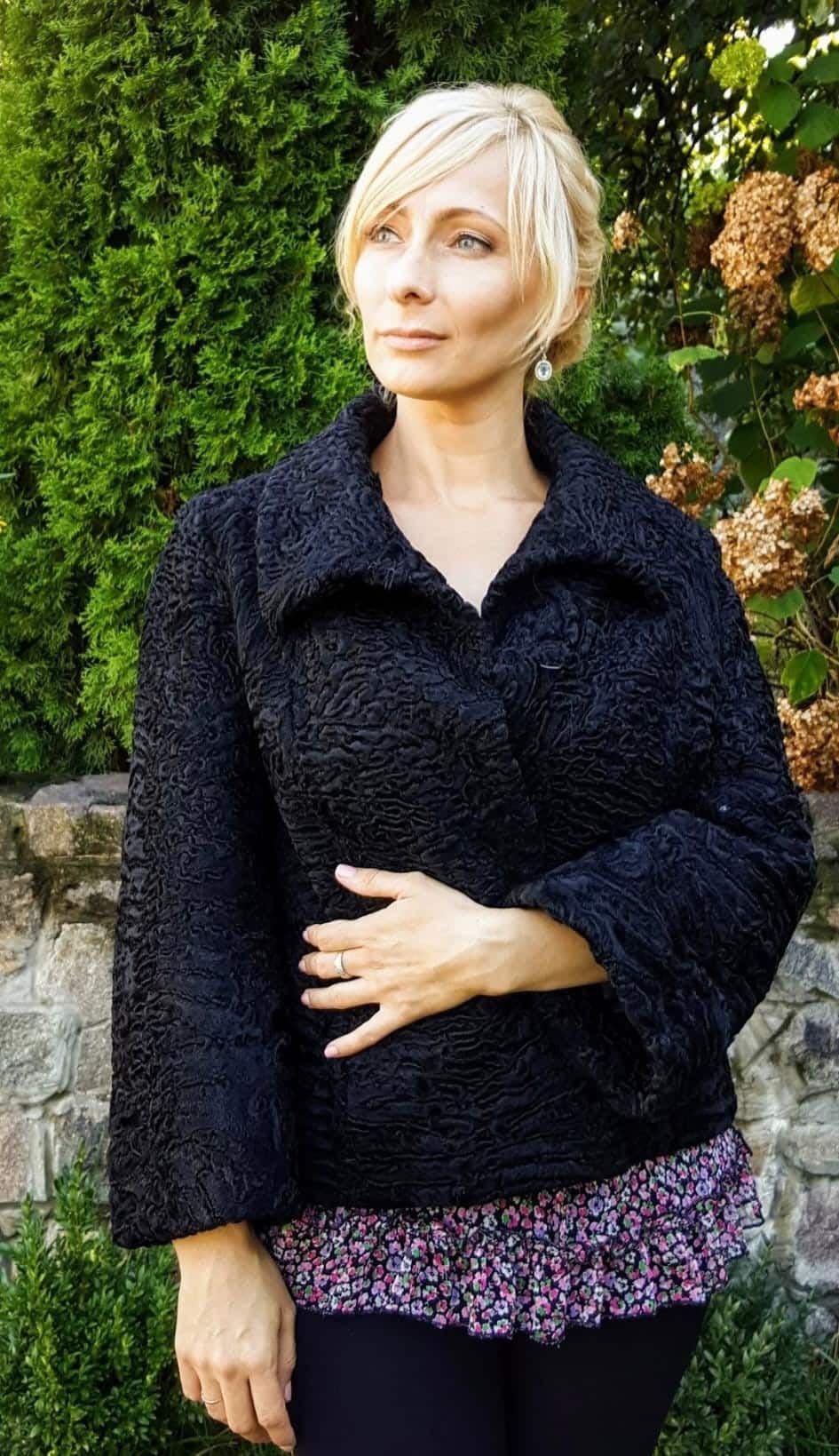 Mujer con chaqueta negra texturizada, mano sobre el estómago, de pie al aire libre.