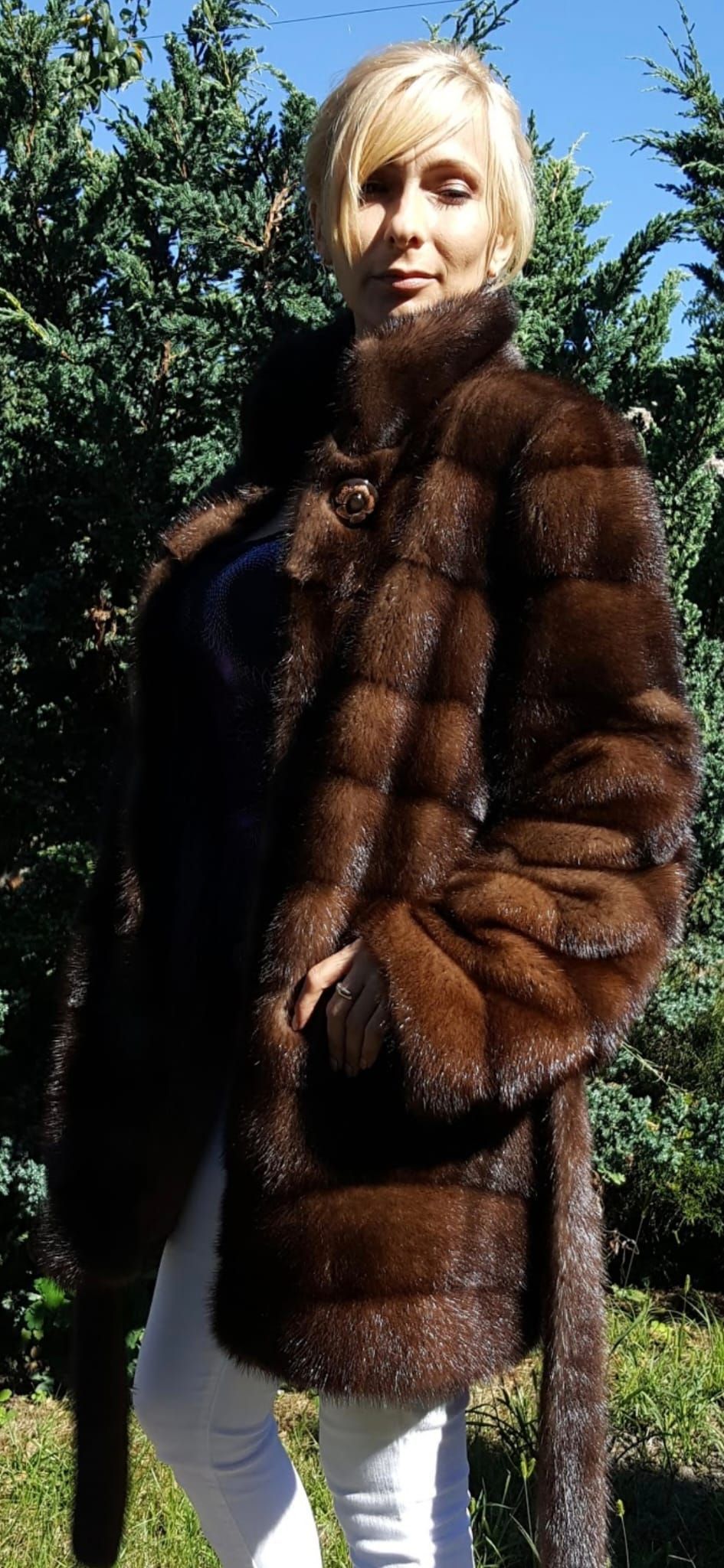 Mujer con abrigo de piel marrón y pantalones blancos, parada afuera.