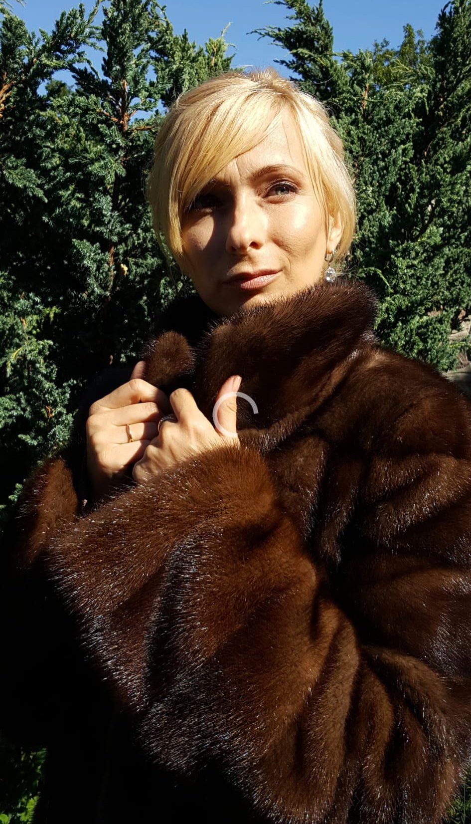 Mujer que lleva un abrigo de piel marrón, sosteniendo el cuello, parada afuera con arbustos verdes en el fondo.