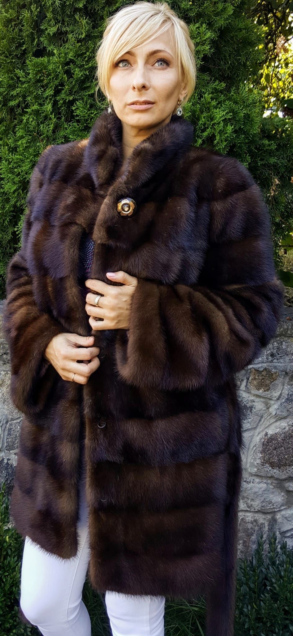 Mujer vestida con un abrigo de piel marrón y pantalones blancos parada afuera, cerca de un muro de piedra.