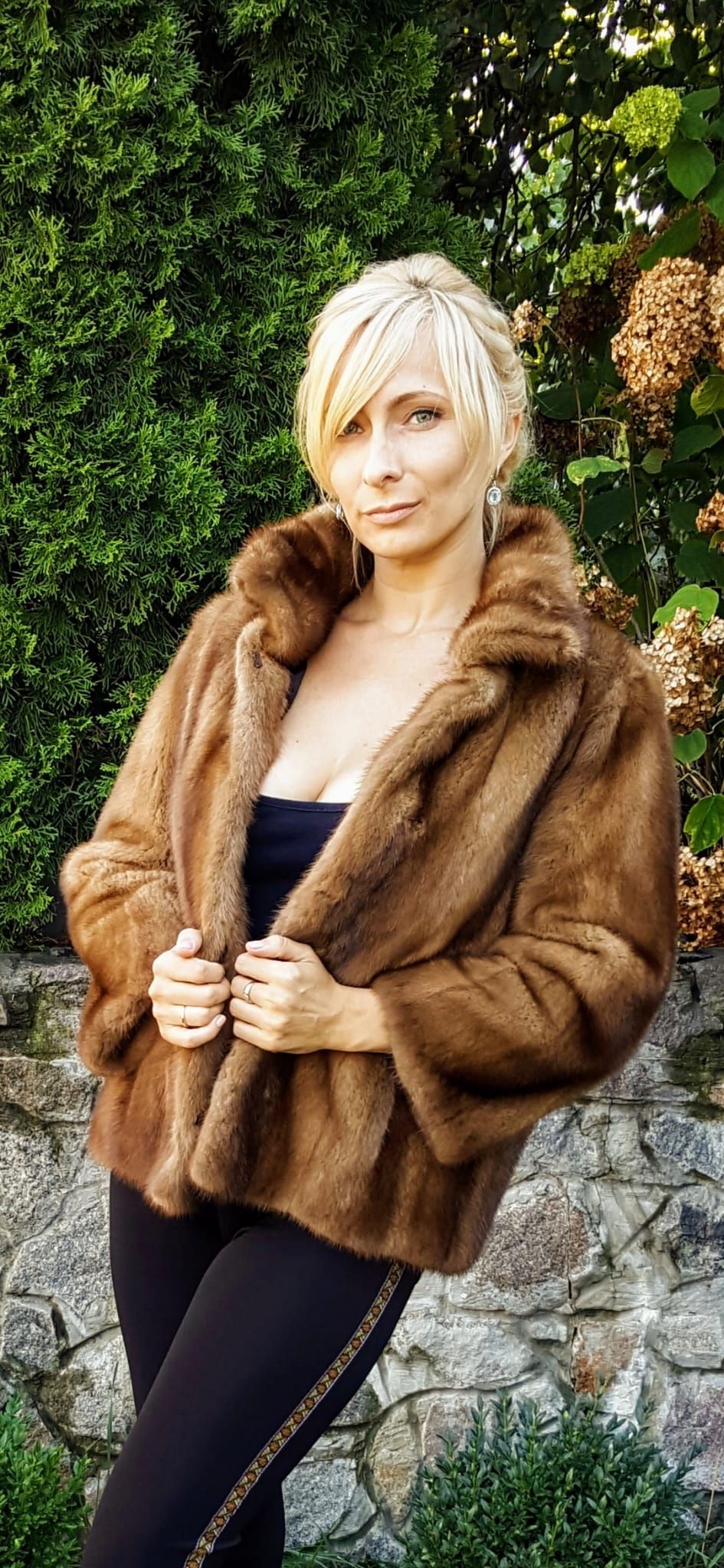 Mujer con un abrigo de piel marrón, un top negro y pantalones, parada afuera.