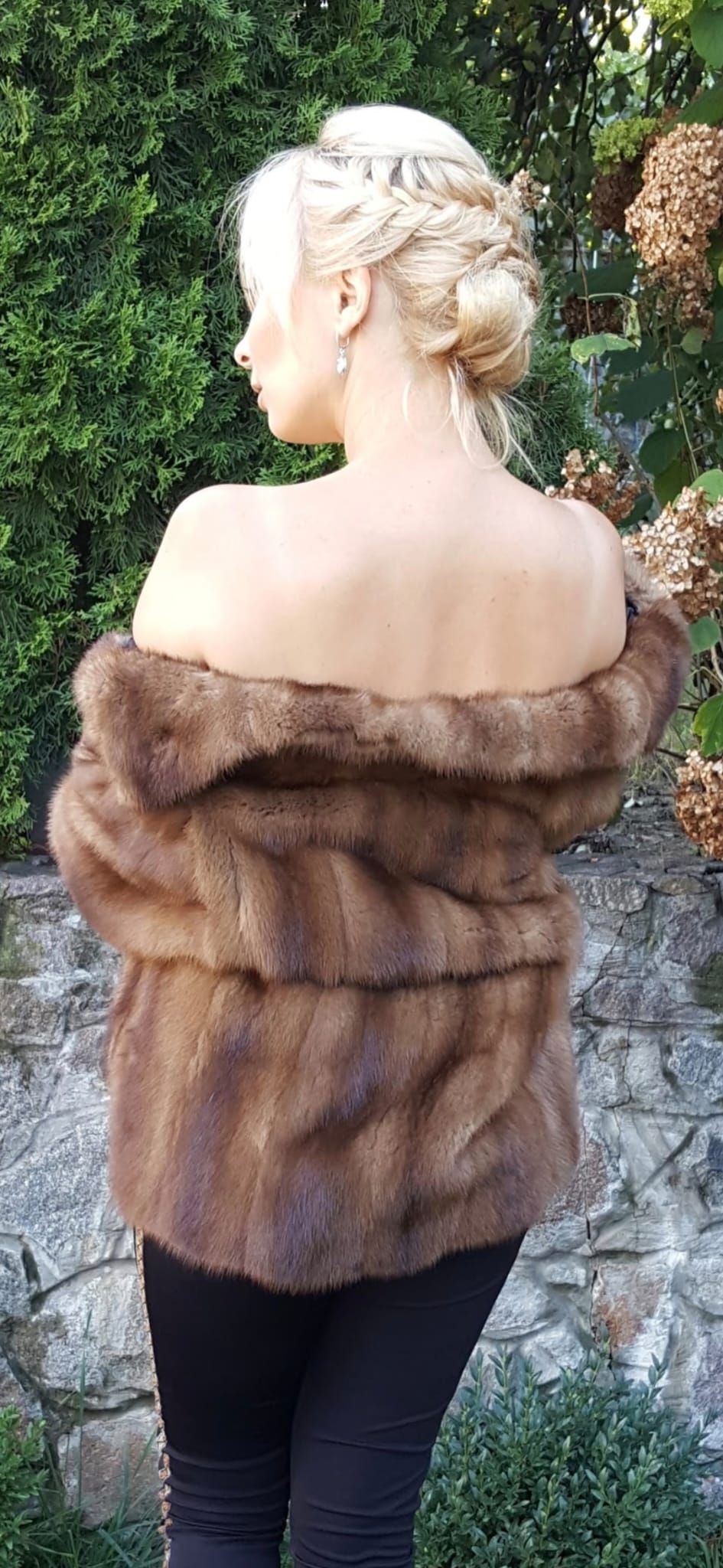 Mujer con una chaqueta de piel marrón, de espaldas a la cámara, de pie al aire libre con follaje verde en el fondo.