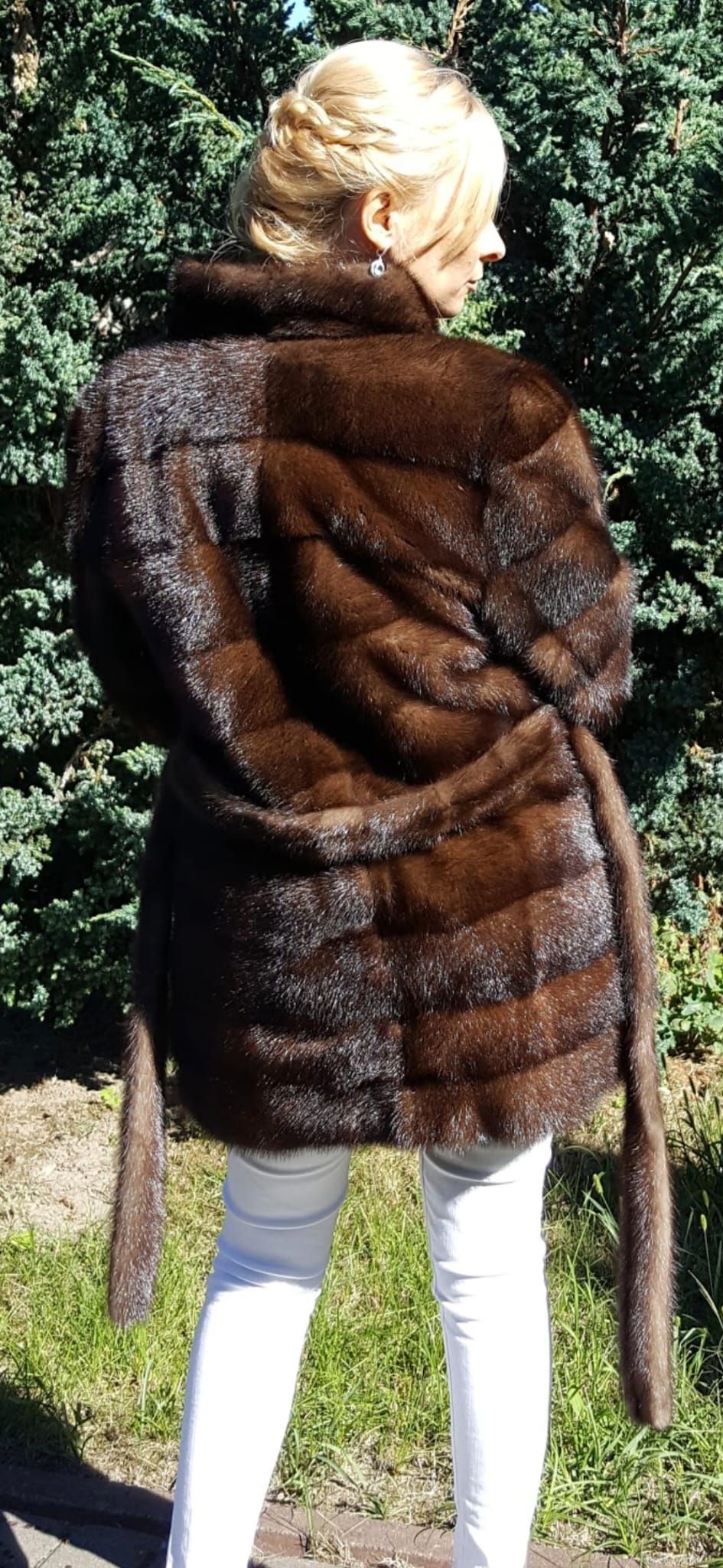 Mujer con un abrigo de piel marrón oscuro, atado a la cintura, y pantalones blancos, de pie al aire libre.