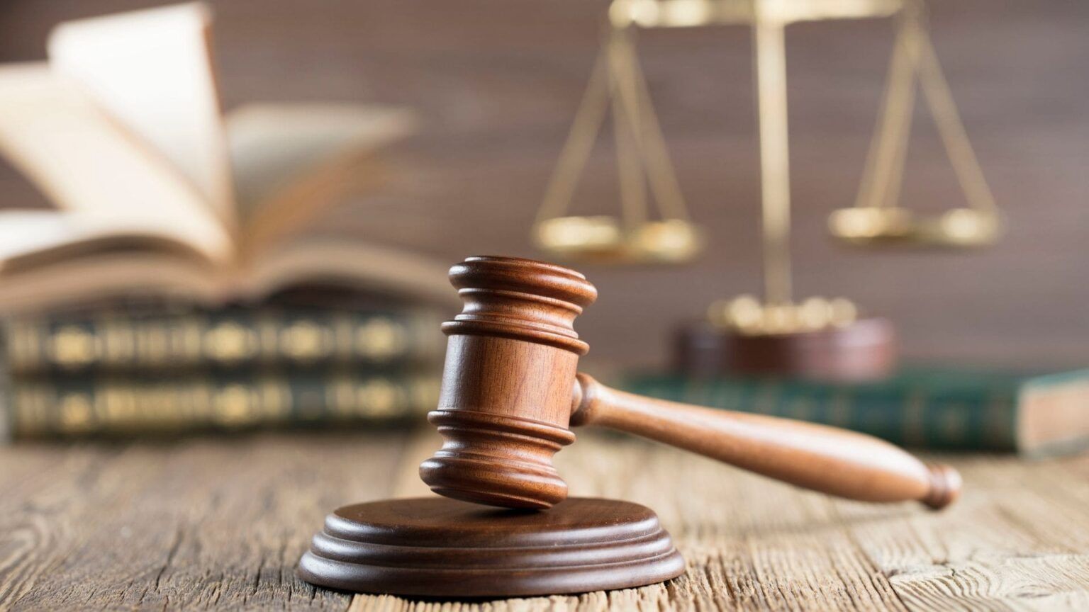 Mazo de juez sobre un escritorio de madera con libros de leyes y balanzas de la justicia al fondo.
