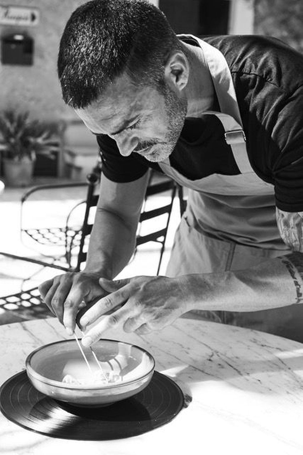 Fotografía en blanco y negro de un hombre concentrado en romper un huevo y echarlo a un bol sobre una mesa. Lleva un delantal al aire libre.