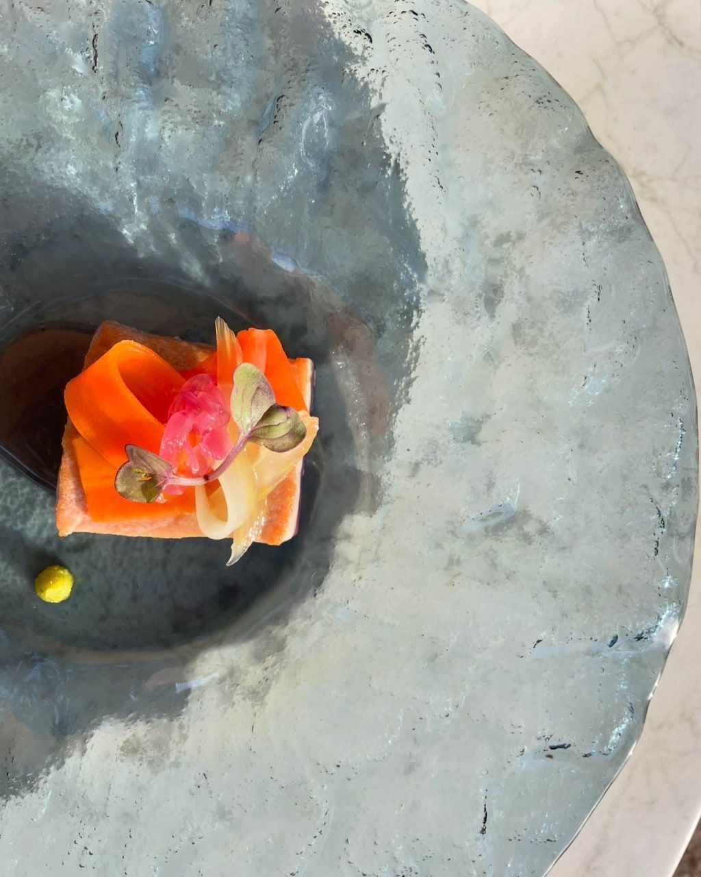 Primer plano de un cuadrado de salmón cubierto con verduras de colores en un plato texturizado de aspecto metálico. Se aprecia un único punto amarillo.