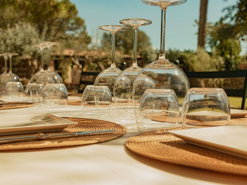 Juego de mesa para exterior con copas de vino, vasos de agua y manteles individuales tejidos, listos para una comida en un día soleado.
