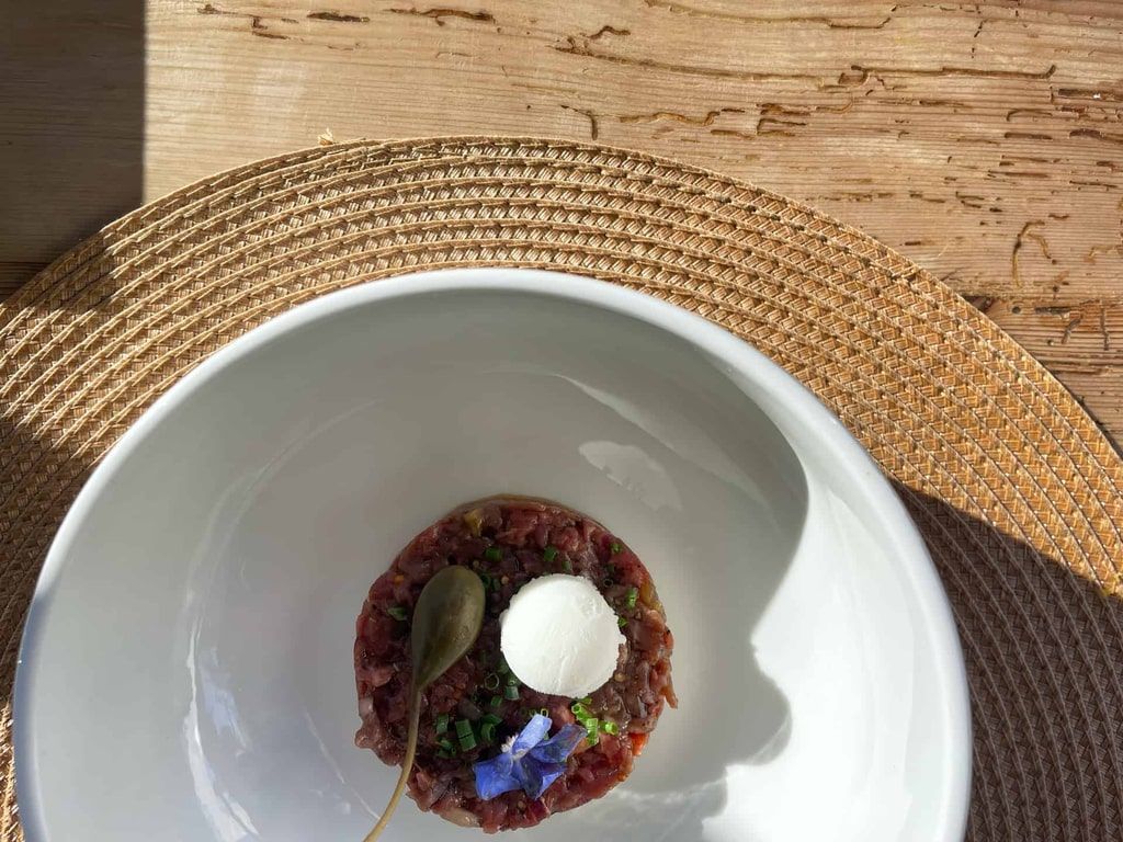 Tartar de ternera en un plato blanco con un mantel redondo de tejido, al aire libre, bajo la luz del sol. Decorado con una cucharada de crema, alcaparras y flores.